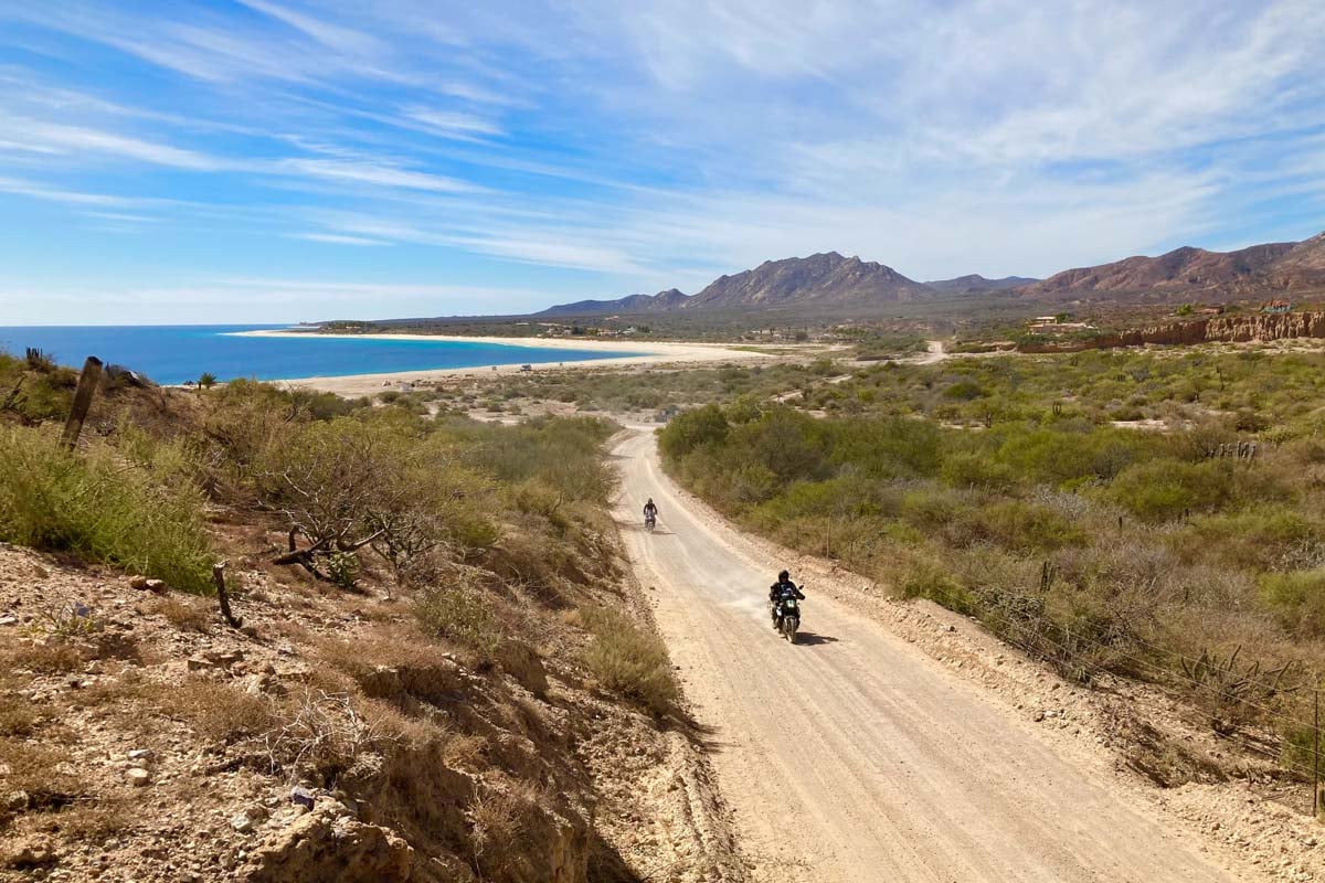 riders-traveling-down-dirt-road-with-ocean-and-mountains-in-background-lars-picture